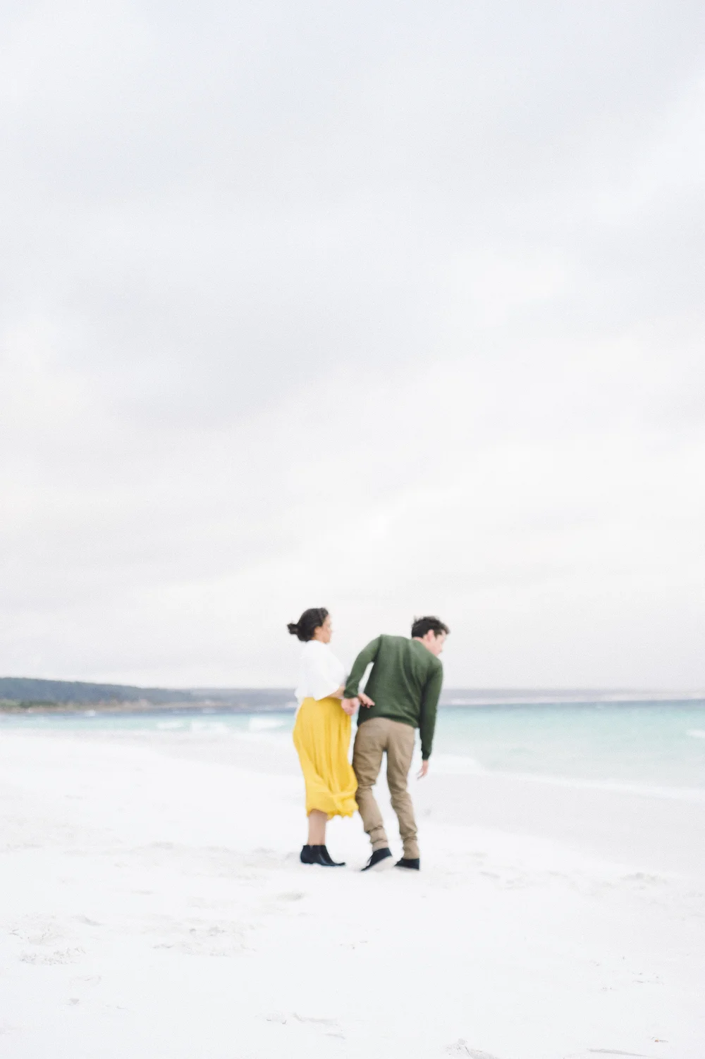 Matte gouache painting of a couple walking along a pale sand shoreline under a soft seafoam sky, warm cream and muted teal tones, tranquil and romantic mood