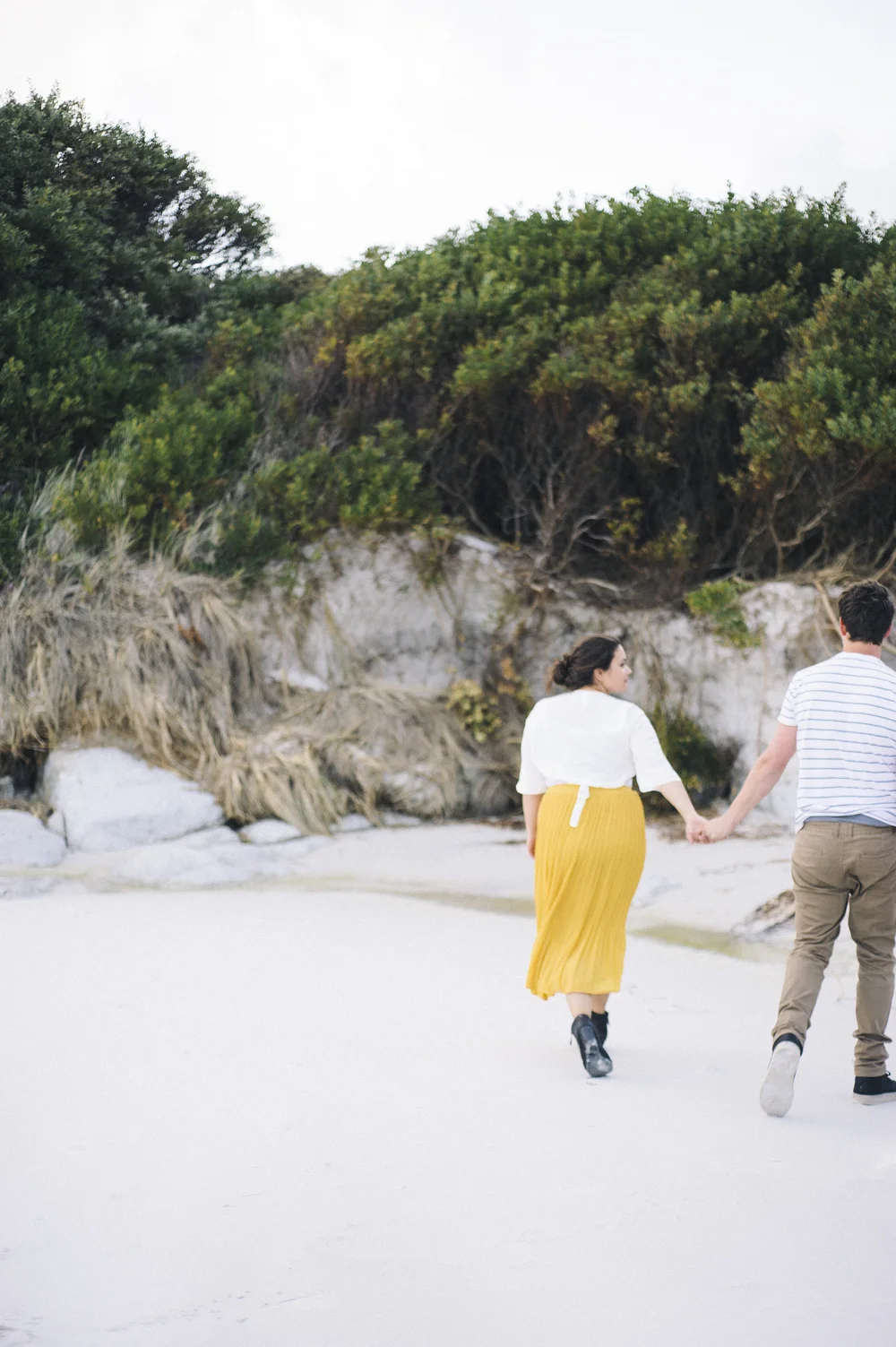 Matte gouache painting of two figures walking toward low coastal shrubs and pale rocks on a sandy beach, soft golden and sage green palette, calm and unhurried mood