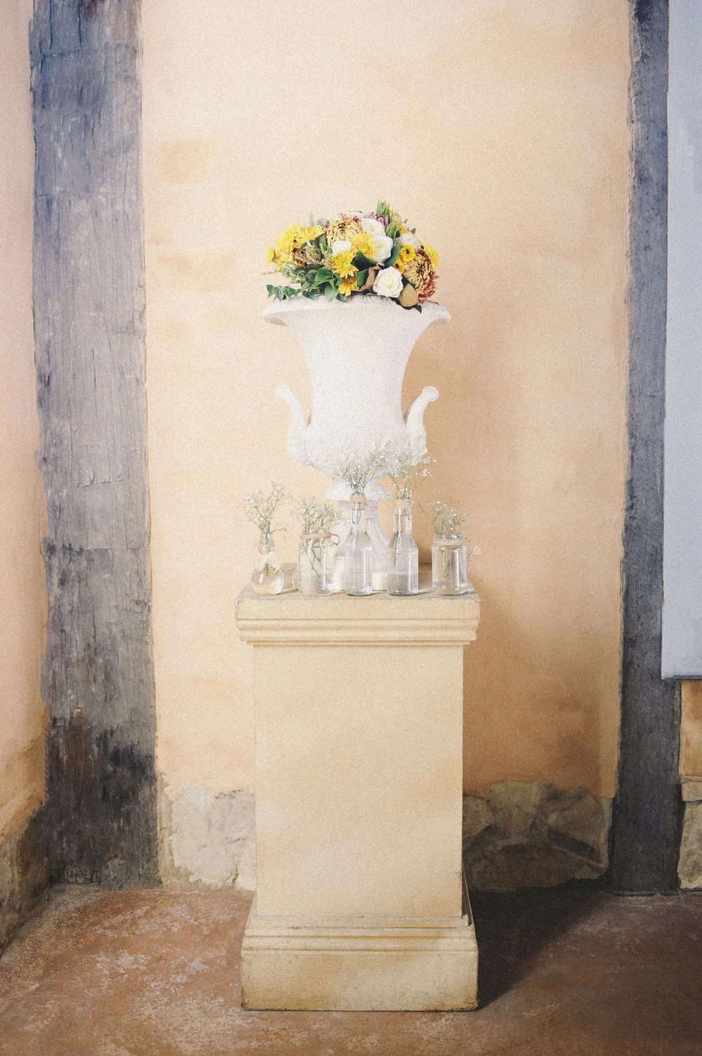 Matte gouache painting of a tall white urn overflowing with cream and golden-yellow flowers on a stone pedestal, surrounded by small glass bottle vases on linen.
