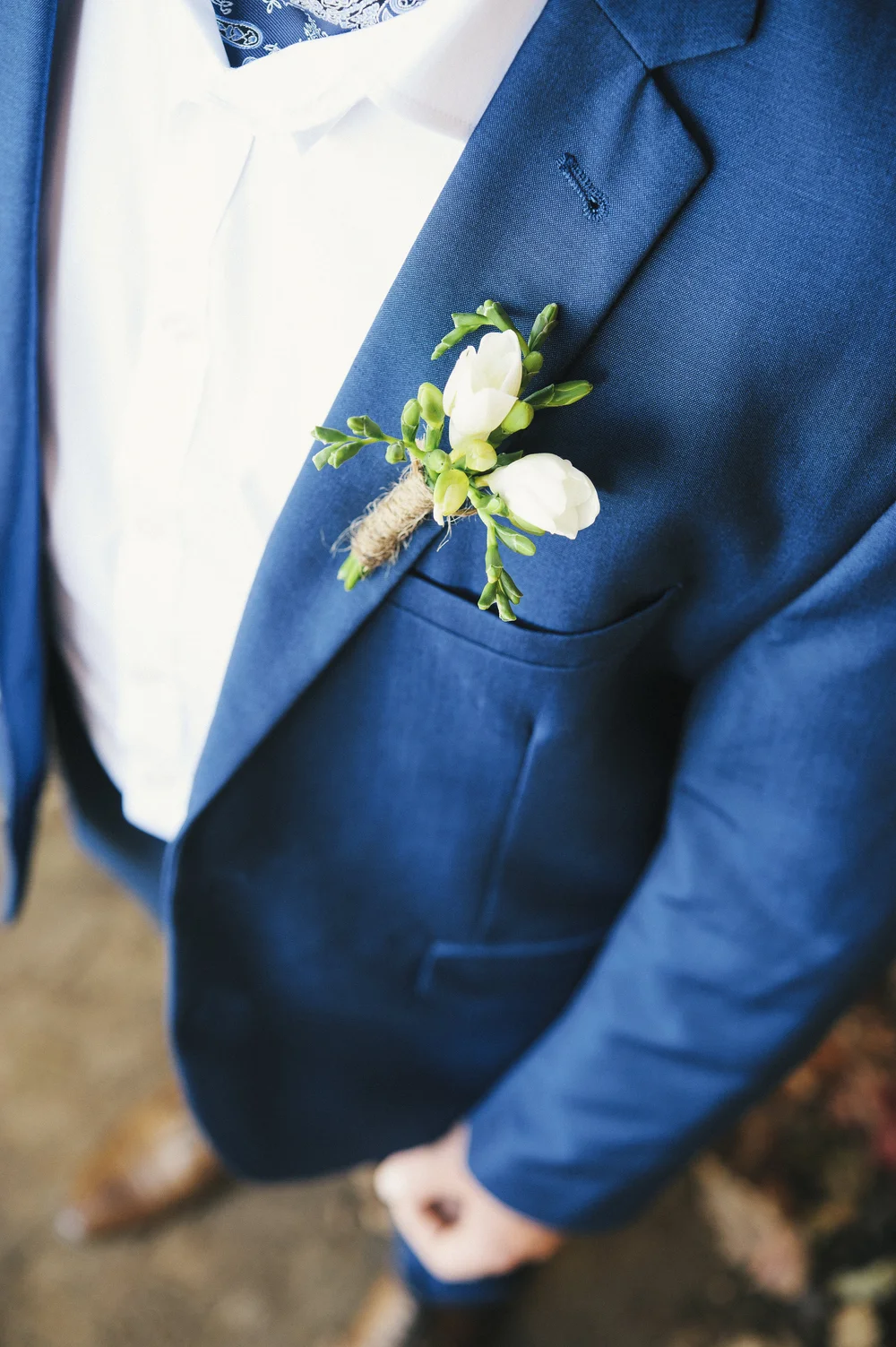 Matte gouache painting of a soft blue suit jacket pocket with a single ivory boutonniere tied with twine, hands clasped in front, earthy greens and creams.